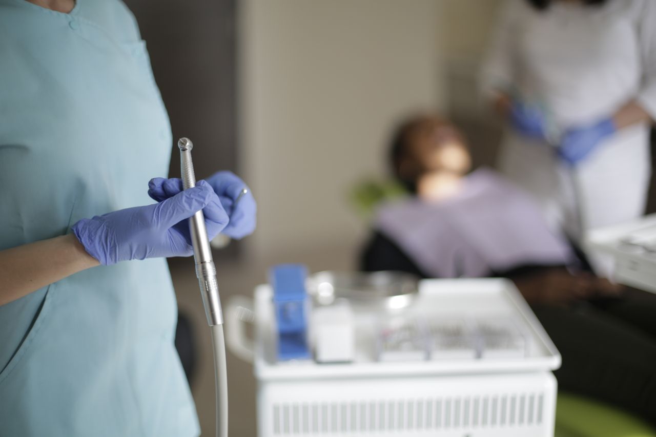 vision Unrecognizable dentist in uniform and gloves standing with dental drill in selective focus while blurred assistant preparing patient for treating teeth
